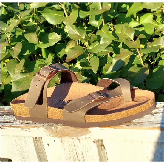 Birkenstock Mayari sandals in Stone Birko-Flor style resting on a wooden surface with green foliage in the background.