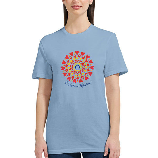Ljusblå figursydd t‑shirt med ett dekorativt cirkelmönster av hjärtan i gult, rött och blått, samt texten ‘Cirkel av Hjärtan’ under motivet.