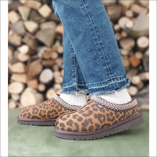 Ugg Tasman Chestnut leopardmönstrade tofflor på fötterna, trendiga och varmfodrade, stående framför en vedstapel.