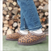 Ugg Tasman Chestnut leopardmönstrade tofflor på fötterna, trendiga och varmfodrade, stående framför en vedstapel.